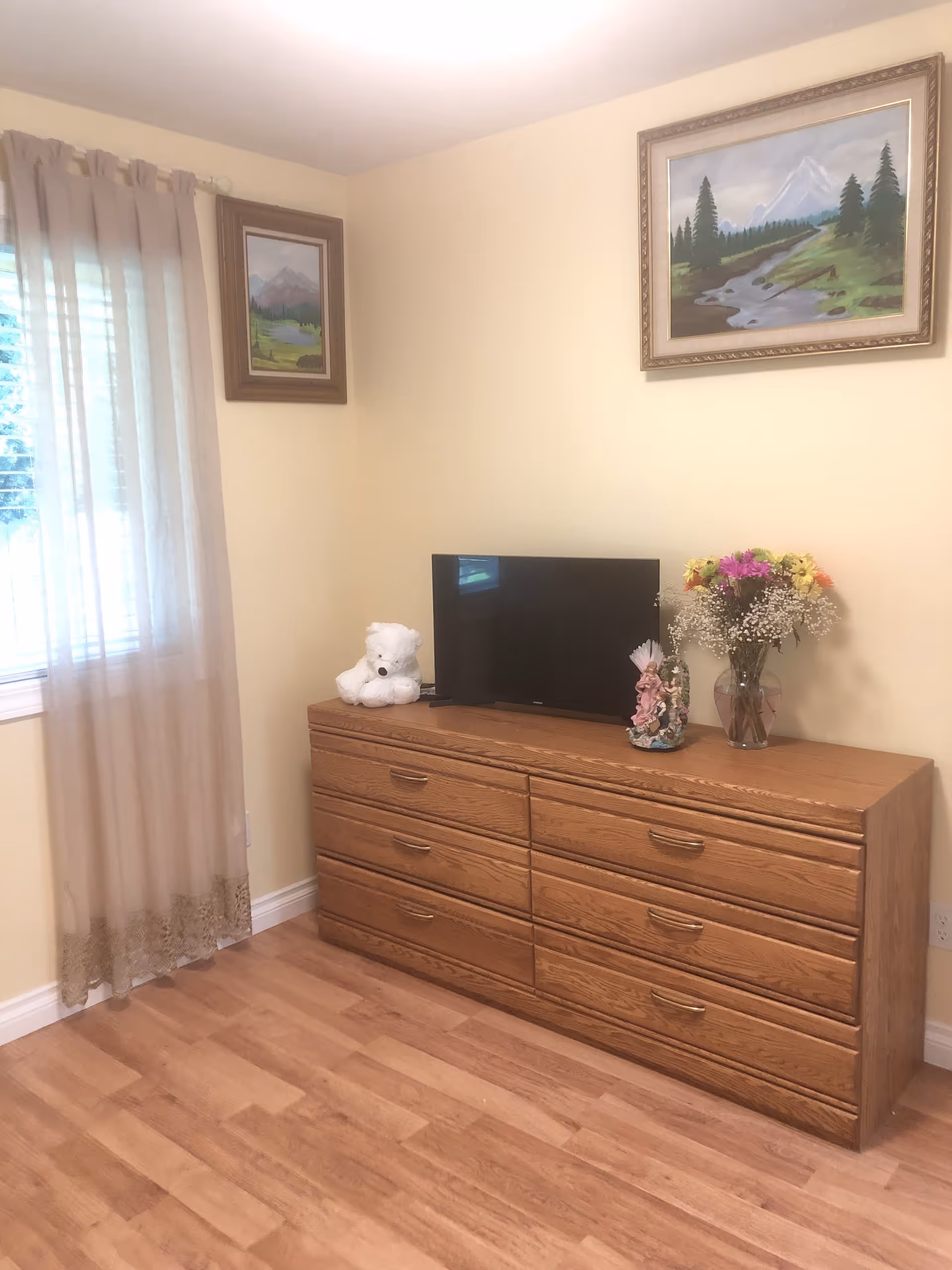 A room with light yellow walls and wooden flooring featuring a wooden dresser with six drawers. On top of the dresser is a flat-screen TV, a white teddy bear, a decorative figurine, and a vase with colorful flowers. Two framed landscape paintings hang on the walls, and a window with sheer beige curtains is on the left side.