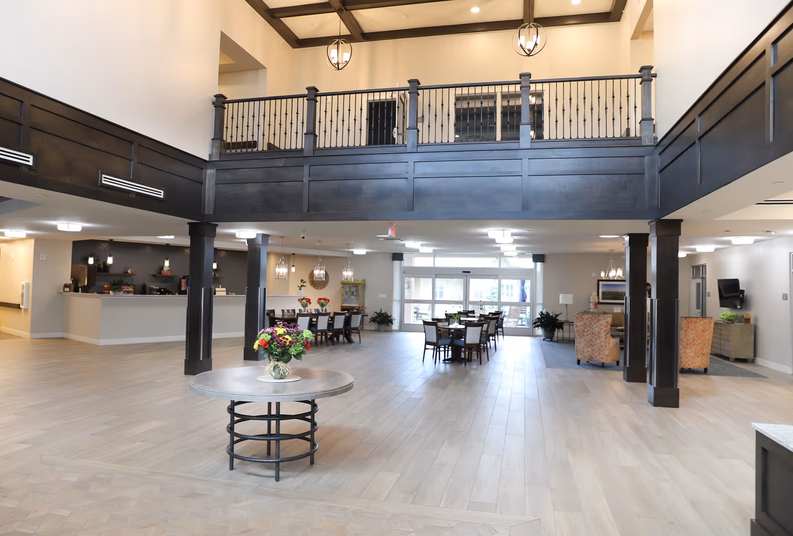 Spacious and well-lit interior of a senior living facility featuring a high ceiling with dark wood balcony railing, a round table with a flower vase in the center, multiple dining tables with chairs, and a seating area with armchairs and a TV on the wall.