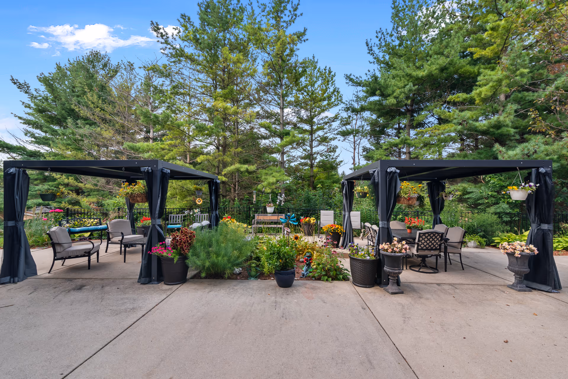 Outdoor patio area with two black pergolas, each furnished with chairs and tables. The space is decorated with various potted plants and flowers, surrounded by lush green trees under a blue sky with some clouds.