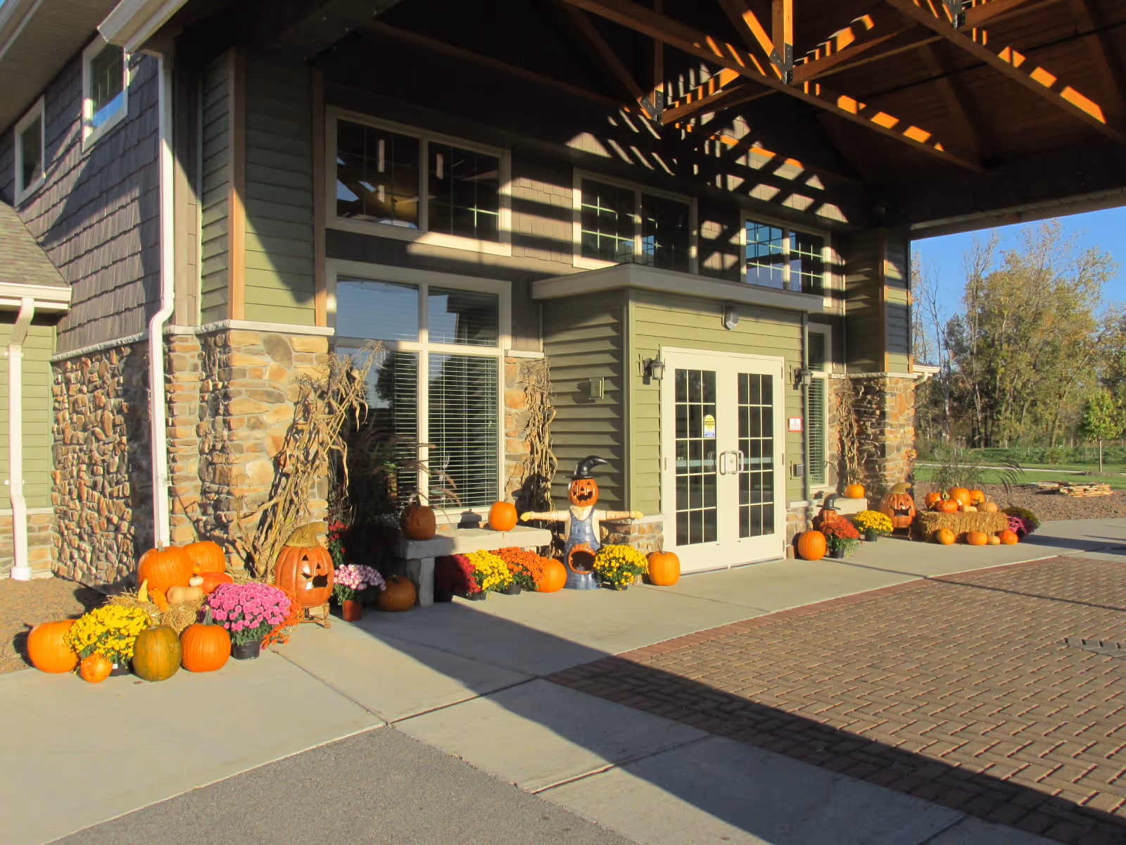 Entrance of a building decorated with pumpkins, colorful flowers, and cornstalks for fall or Halloween. The building has stone and green siding with large windows and double glass doors under a wooden overhang.