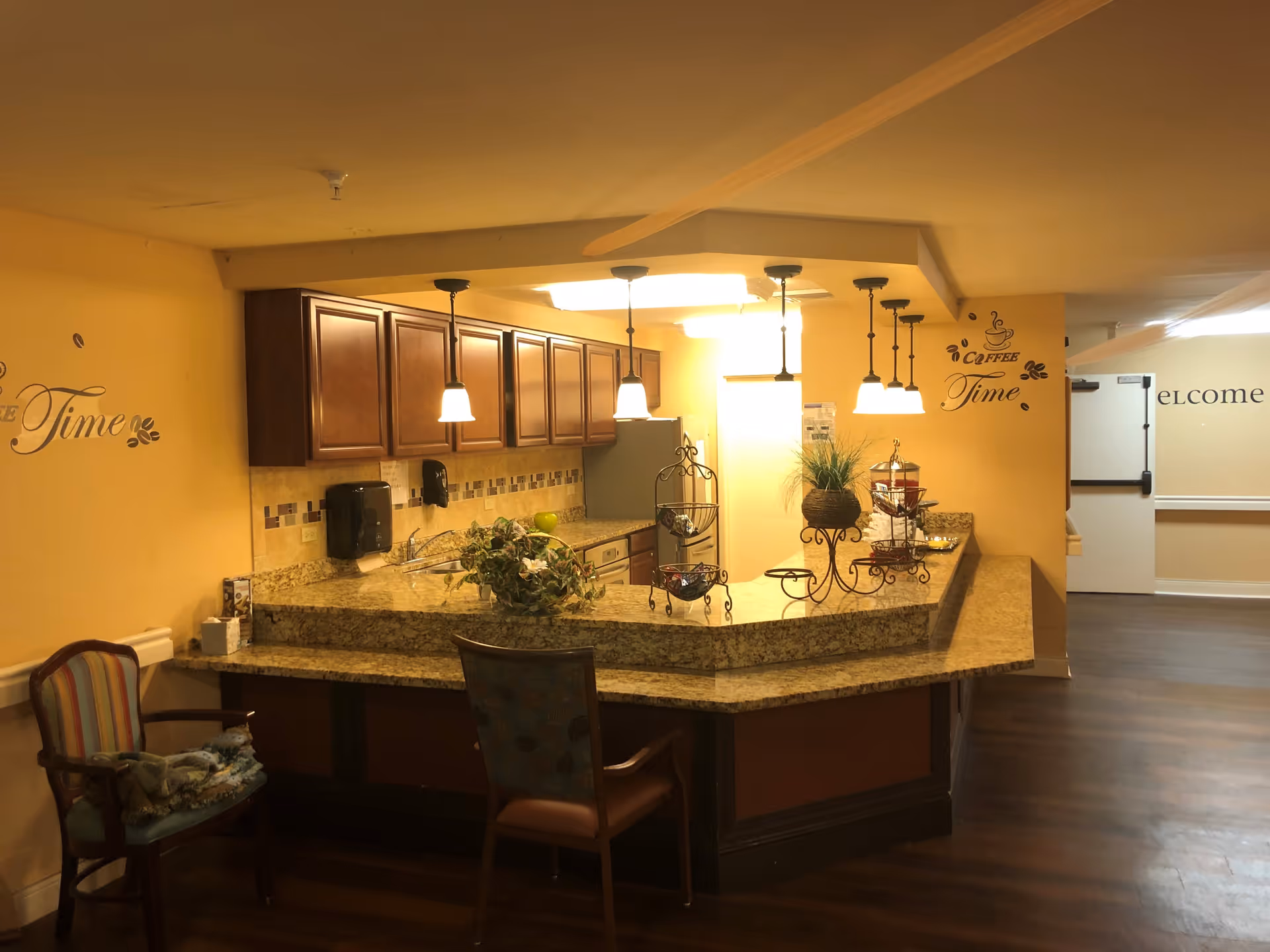 Interior view of a kitchen area with a granite countertop bar, wooden cabinets, and hanging pendant lights. There are decorative plants and a tiered stand on the counter. Two chairs are placed near the counter, and the walls are painted yellow with wall decals that say 'Coffee Time'. A hallway with a door is visible in the background.