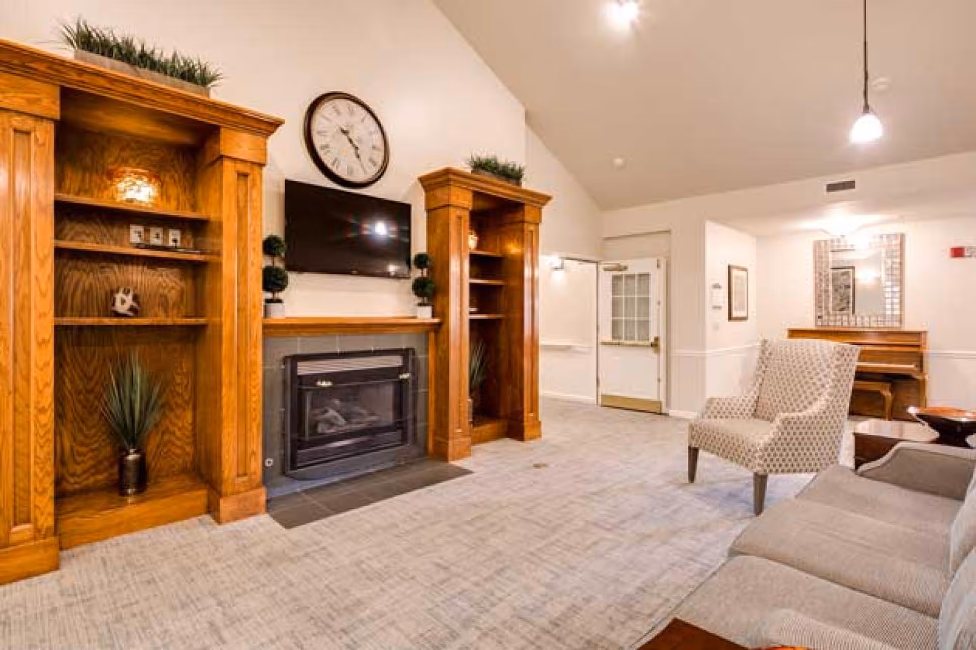 Bright common living room with built-in wooden shelves and a fireplace, wall-mounted TV, and upholstered seating.