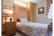 Small bedroom with a single bed dressed in patterned bedding, a wooden nightstand with a lamp, framed art on the wall, and a window with blinds.