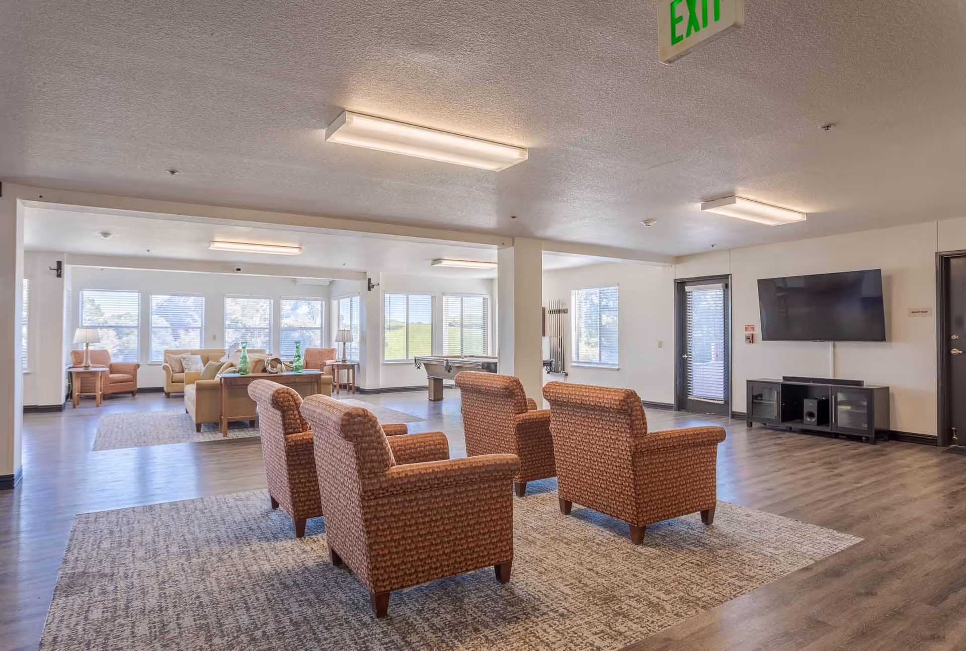 A spacious senior living facility common area with multiple seating arrangements including four patterned armchairs on a rug in the foreground and sofas with side tables and lamps near large windows in the background. A flat-screen TV is mounted on the wall above a media console, and a pool table is visible near the windows. The room has wood flooring and ceiling lights, with an exit sign overhead.