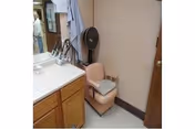 Bathroom vanity area with a sink and wooden cabinet, a padded beige chair beside a standing fan and hanging towels.