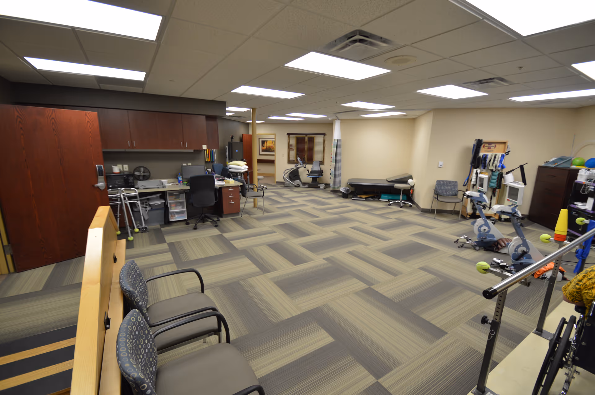 A spacious therapy or rehabilitation room with exercise equipment including stationary bikes, parallel bars, and therapy tables. There are chairs along the wall, a desk with office supplies, and storage cabinets. The room has a patterned carpet and bright overhead lighting.