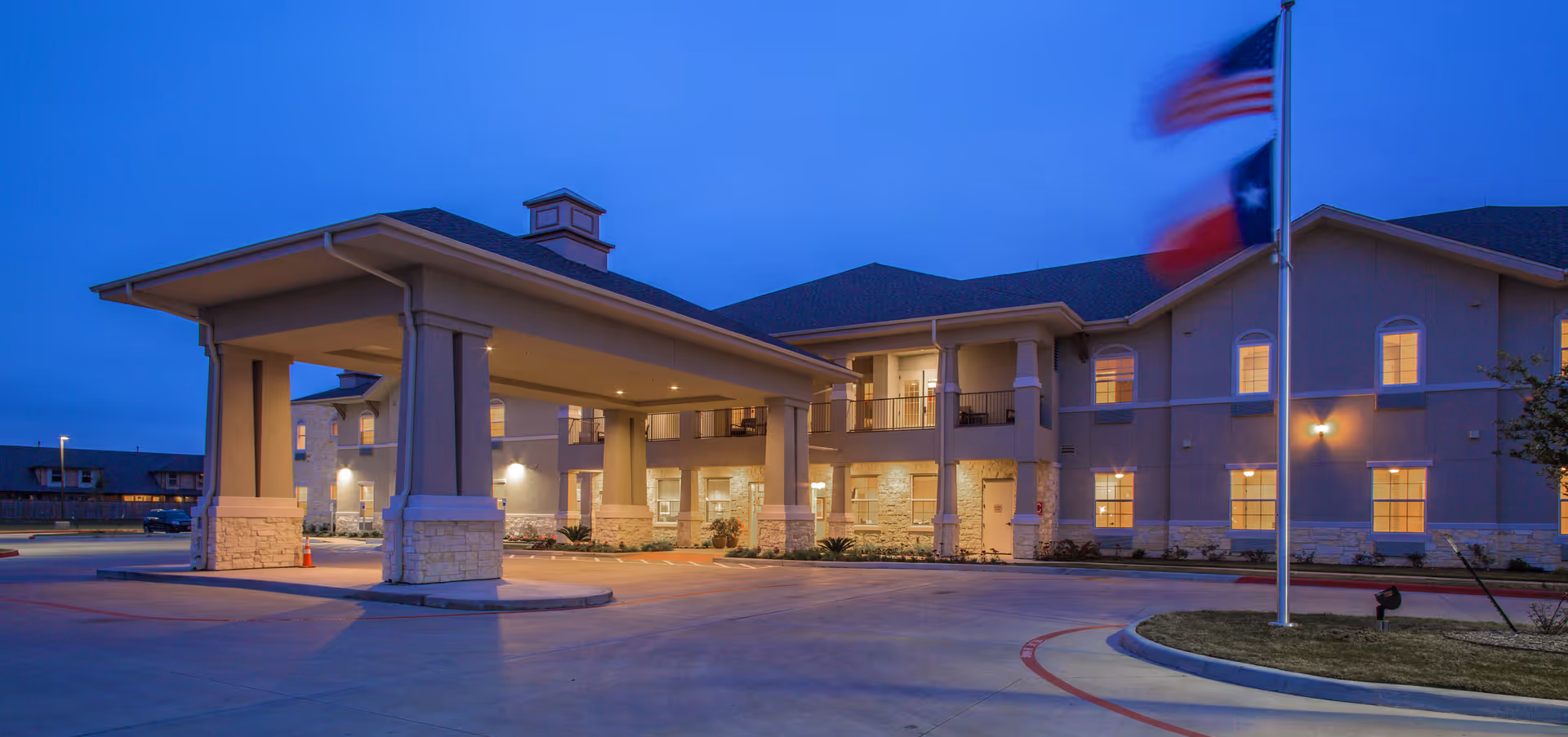 Exterior view of The Legacy at Falcon Point senior living facility at dusk, showing a covered entrance with pillars, illuminated windows, and an American flag and Texas state flag on a flagpole in the foreground.