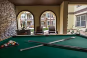 A covered outdoor seating area with a pool table in the foreground. Two pool cues are resting on the green felt surface of the table, with a rack of billiard balls at one end. Beyond the arches of the covered area, there are two chairs and a view of a courtyard with plants and a multi-story building in the background.