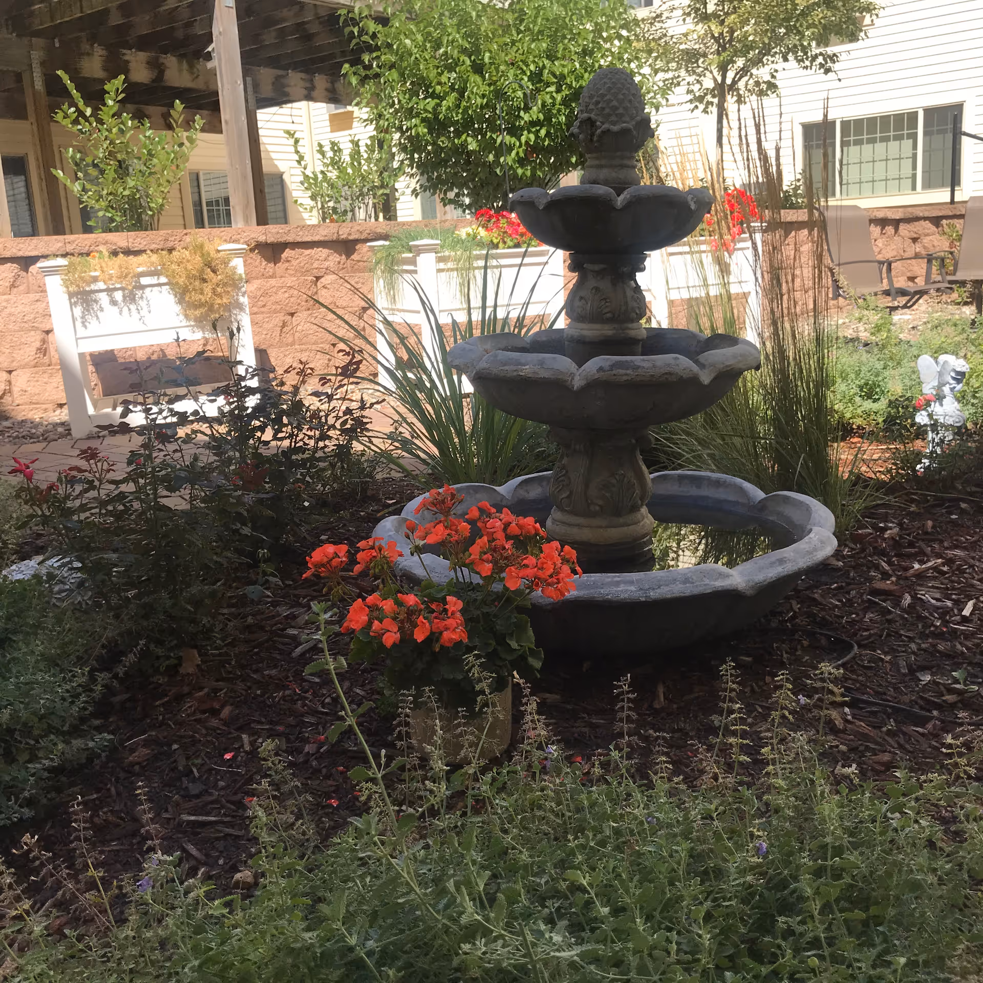 Outdoor garden area with a three-tier stone fountain surrounded by various plants and flowers, including bright red blooms. In the background, there is a stone wall, some patio chairs, and part of a building with windows.