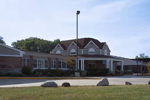 Exterior view of a single-story senior living facility building with a covered entrance, brick and siding facade, surrounded by greenery and a clear sky.