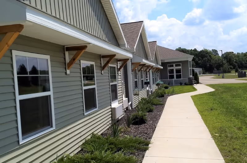 Exterior view of a single-story building with multiple windows and a sloped roof. A concrete walkway curves alongside the building, bordered by landscaped plants and grass. Trees and a partly cloudy sky are visible in the background.