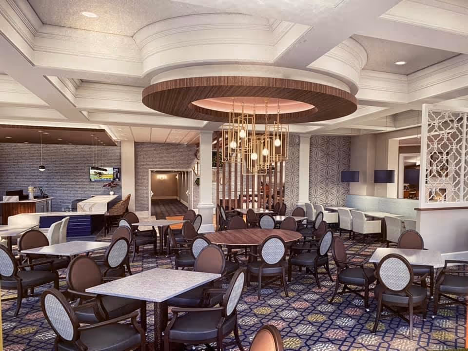 A spacious and elegantly decorated dining area with multiple tables and chairs arranged neatly. The room features a patterned carpet, a modern chandelier with multiple hanging lights, and a coffered ceiling with recessed lighting. There are decorative wall panels and a small reception or service desk area in the background.