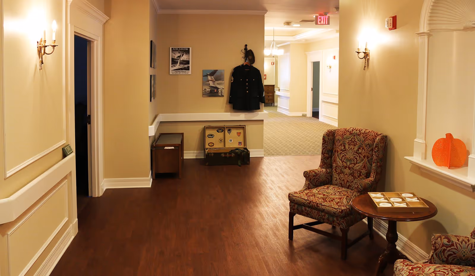 A hallway in a senior living facility with beige walls and wooden flooring. There are two patterned armchairs and a small round table with a tic-tac-toe game on it. On the right wall, there is a decorative orange pumpkin. At the end of the hallway, a military jacket and hat are hanging on the wall above a vintage suitcase. Wall sconces provide warm lighting, and there are framed pictures on the walls. The hallway leads to a carpeted area with more rooms and an exit sign visible.