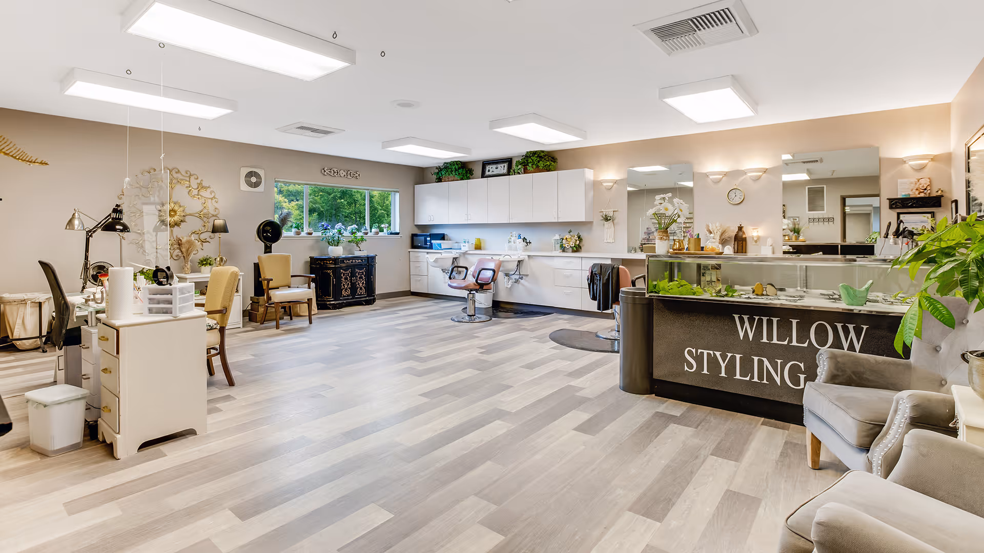 A spacious and well-lit styling salon with light wood flooring, white cabinetry, and multiple styling stations including chairs and mirrors. There is a reception desk with the words 'WILLOW STYLING' on the front, comfortable seating, decorative plants, and a window showing greenery outside.