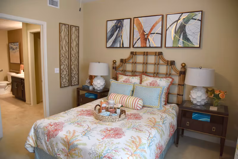 A cozy bedroom with a bed featuring a coral and seaweed patterned bedspread and multiple pillows. The bed has a wooden headboard and is flanked by two dark wood nightstands, each with a white lamp. Above the bed are three abstract paintings with curved lines in various colors. To the left, an open door reveals a bathroom with a toilet and sink visible.