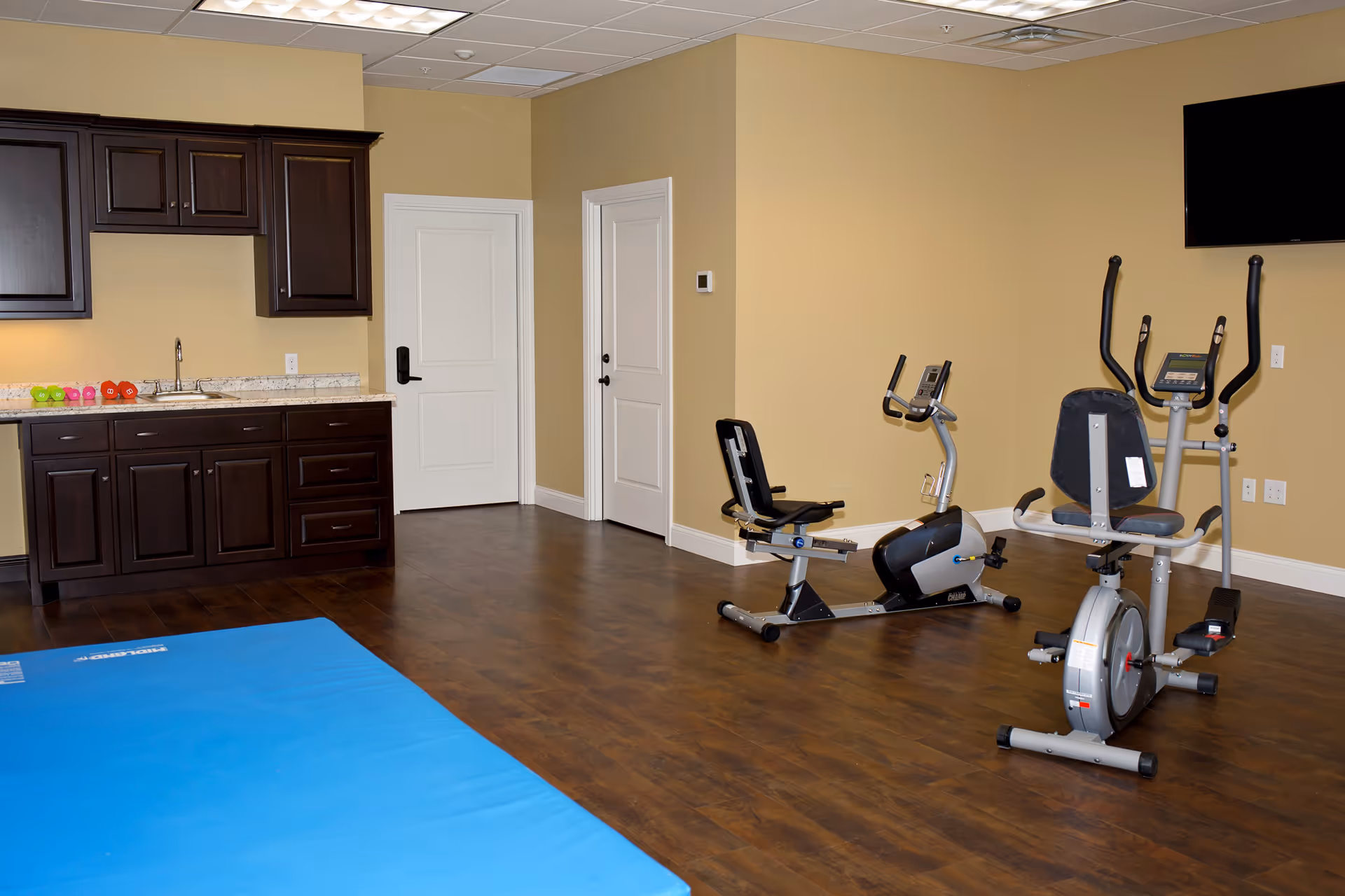 Fitness room with two recumbent exercise bikes, a blue exercise mat, and dark wood cabinets along one wall.