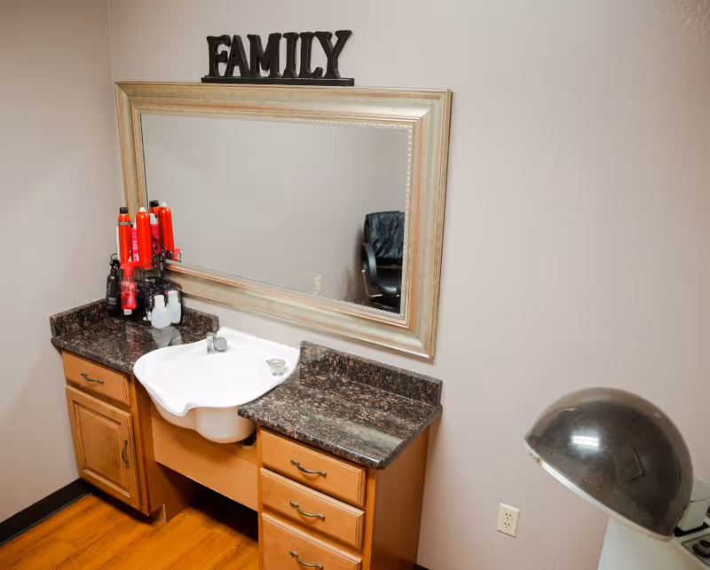 A vanity area with a large rectangular mirror framed in light wood, a white sink set into a dark granite countertop with wooden cabinets below. On the left side of the countertop are several red and black hair care products. Above the mirror is a decorative sign that reads 'FAMILY'. To the right is a vintage hair dryer chair with a dome-shaped hood dryer.