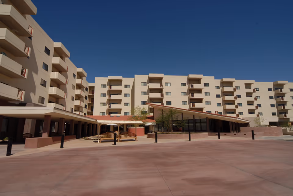 Exterior view of Nunnenkamp Center Friendship Village Tempe, showing a multi-story beige building with balconies under a clear blue sky. The foreground features a large paved area and a covered entrance with benches and some small trees.