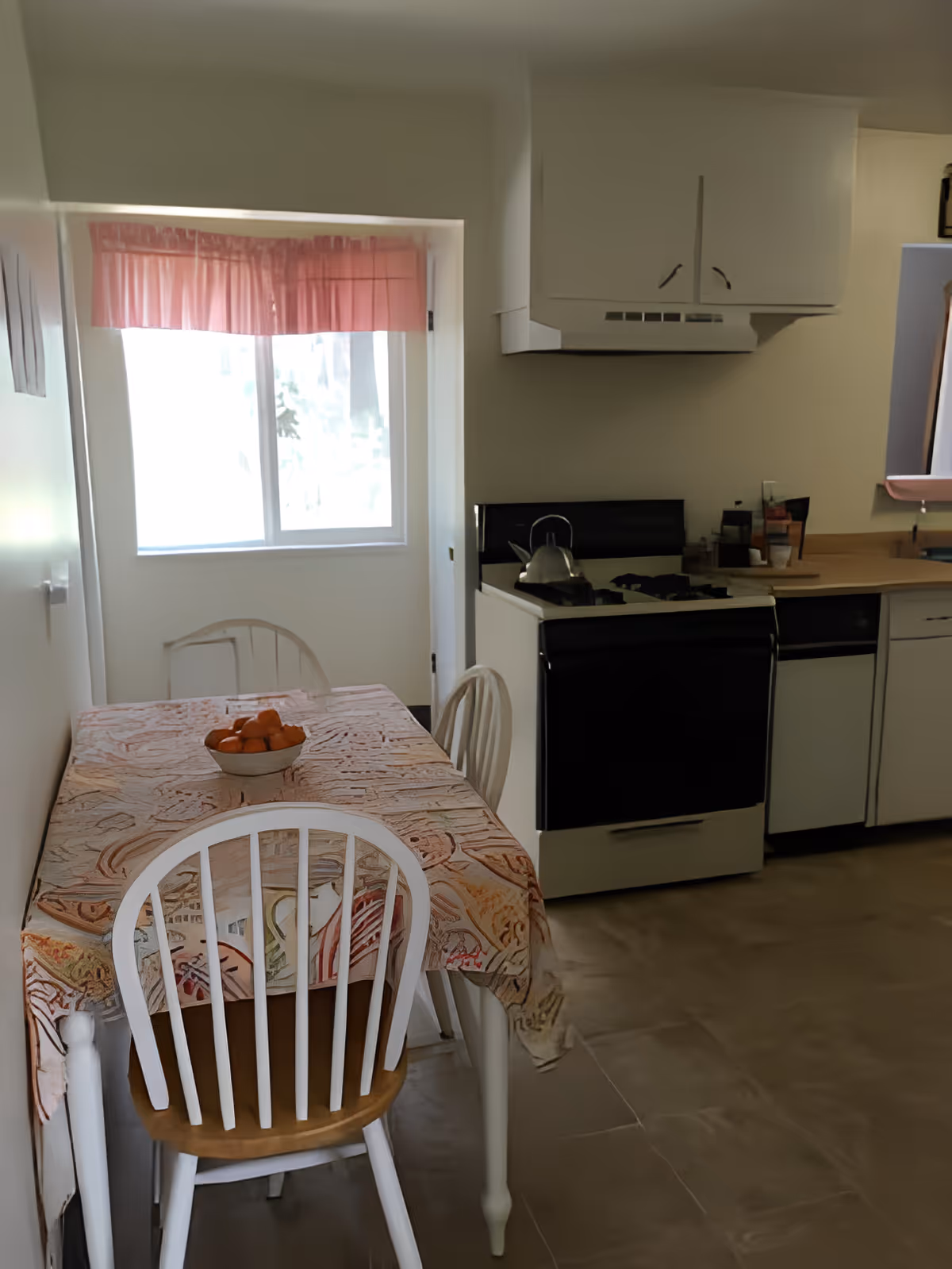 A small kitchen and dining area with a table covered by a patterned tablecloth and three white wooden chairs. A bowl of oranges is placed on the table. The kitchen features a white stove with a kettle on it, white cabinets, and a window with pink curtains letting in natural light.