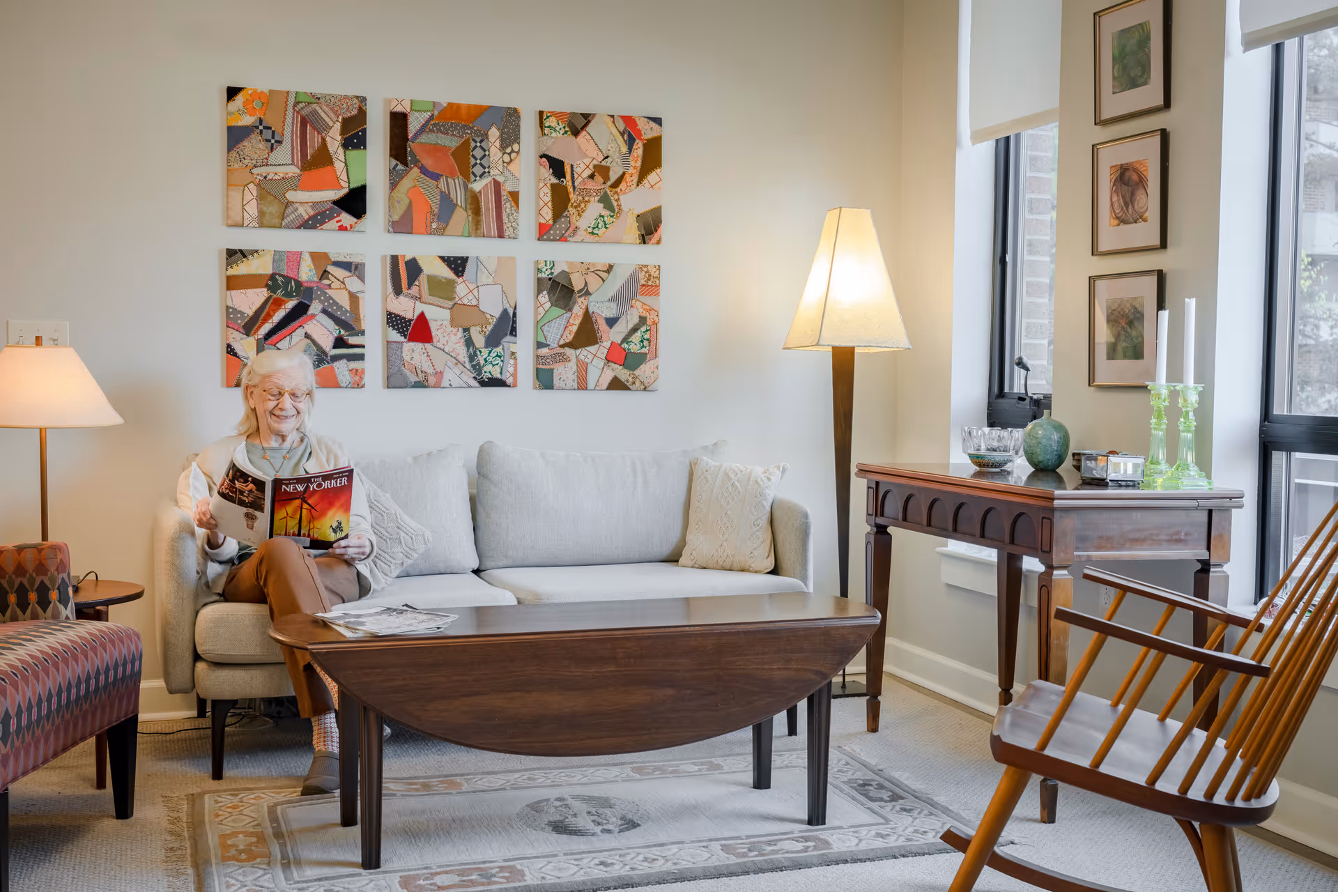 An elderly woman sitting on a light gray sofa in a cozy living room, reading The New Yorker magazine. The room features a wooden coffee table, a patterned armchair, a floor lamp, and a wooden side table with decorative items. Six colorful abstract art pieces hang on the wall above the sofa, and three framed pictures are displayed near a window with a view outside.