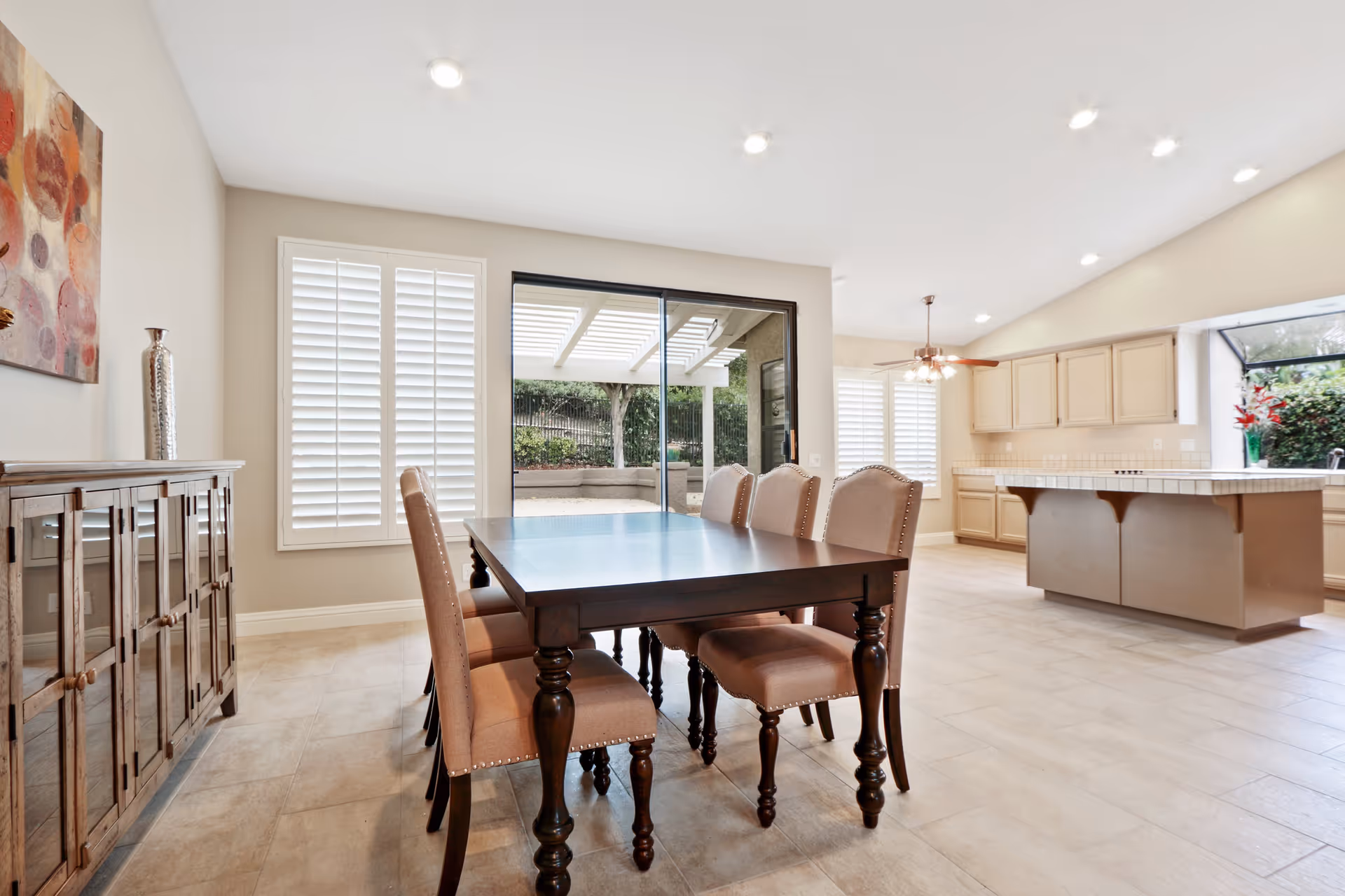 Bright dining area with a dark wooden table and six upholstered chairs. The room features large windows with white shutters, a sliding glass door leading to an outdoor patio, and a kitchen with beige cabinets and a large island in the background.