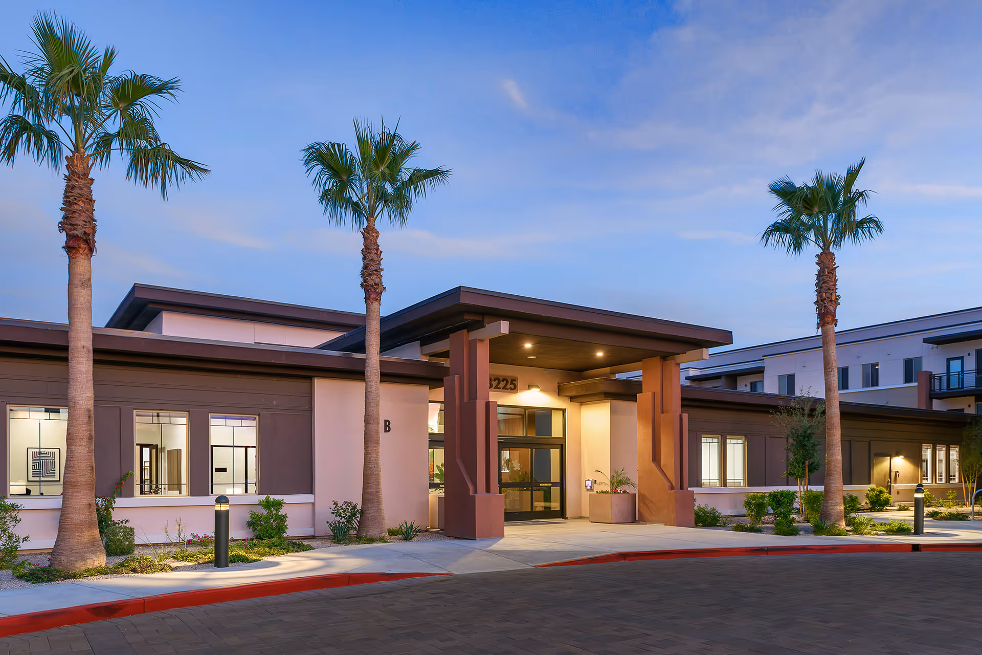 Exterior view of a modern senior living facility building at dusk with palm trees and well-lit entrance under a covered portico.