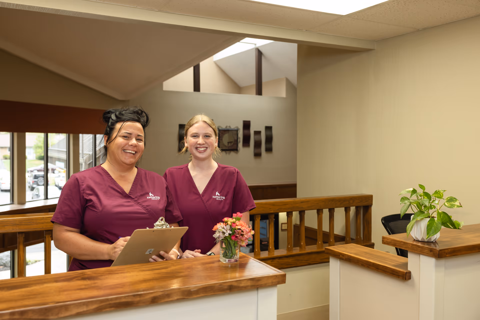 Two smiling healthcare workers wearing maroon scrubs stand behind a wooden reception desk in a well-lit interior space. One holds a clipboard, and there is a small vase with flowers on the desk. A potted plant is on a nearby counter, and large windows and decorative wall art are visible in the background.