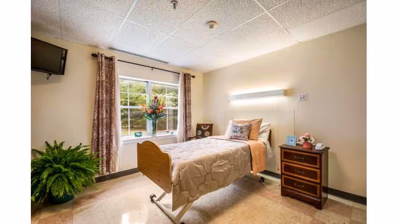A single bed in a nursing center room with beige bedding and pillows, a wooden nightstand with a telephone, a small framed photo, and a flower vase. There is a window with floral curtains letting in natural light, a green potted plant on the floor, and a wall-mounted TV in the corner.