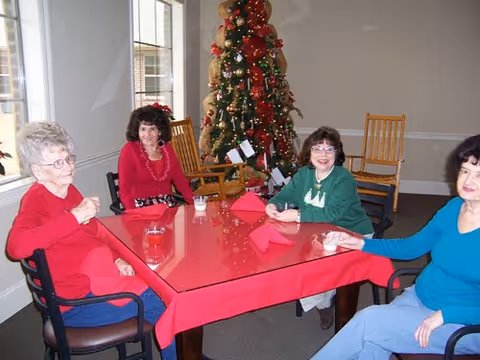 Four elderly women sitting around a rectangular table covered with a red tablecloth in a room decorated with a Christmas tree in the background. The women are smiling and appear to be enjoying each other's company.