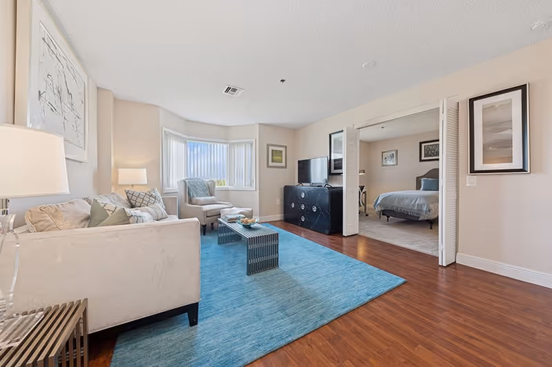 A bright and spacious living room with a beige sofa, armchair, and a blue area rug on wooden flooring. A coffee table is centered on the rug, and a TV sits on a black cabinet against the wall. The room opens into a bedroom with a bed and framed artwork on the walls. Large windows with vertical blinds allow natural light to fill the space.