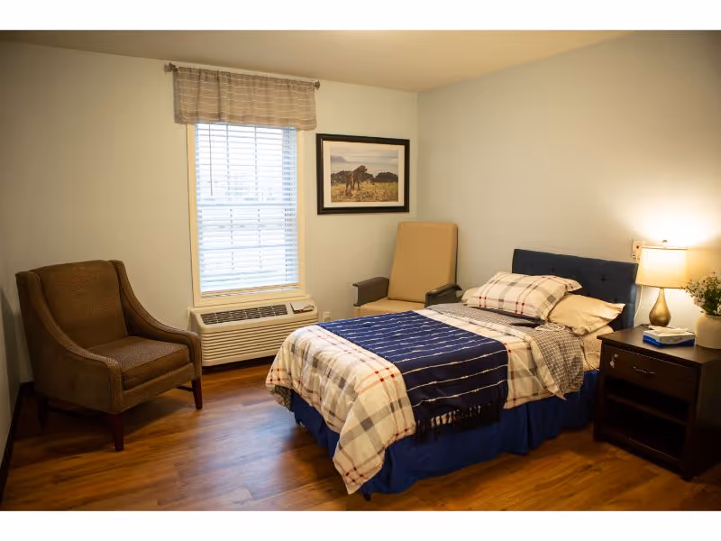 A cozy bedroom with a single bed covered in plaid bedding and a blue throw blanket. There is a brown armchair to the left, a beige armchair near the corner, and a nightstand with a lamp and a plant on the right side of the bed. A window with blinds and a valance is centered on the wall behind the bed, and a framed picture hangs above the beige chair. The room has wooden flooring and light-colored walls.