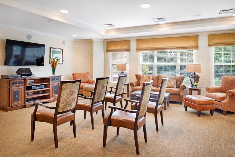 A well-lit living room with six wooden chairs arranged in two rows facing a wall-mounted flat-screen TV. The room features a wooden media console with a vase of flowers and framed artwork on the wall. There are large windows with beige blinds, a brown sofa with floral cushions, two armchairs, a matching ottoman, and two side tables with lamps.