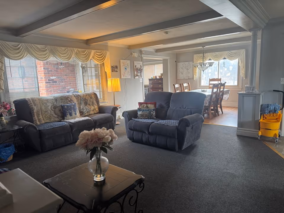 A cozy living room with two dark gray recliner sofas adorned with patterned pillows and a throw blanket. A small table with a vase of flowers is in the foreground. The room has large windows with white curtains and a standing lamp. In the background, there is a dining area with a wooden table and chairs, a chandelier, and a window with similar curtains. A yellow cleaning cart is visible near the dining area.