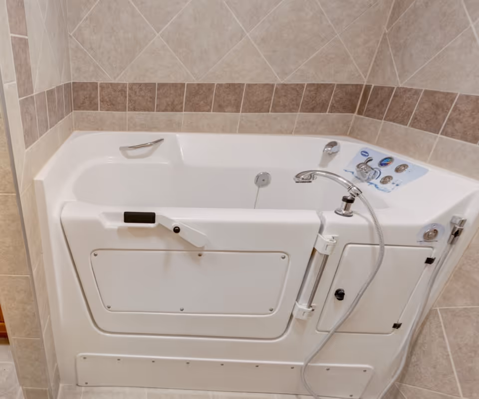 Walk-in bathtub with a hinged door, handheld showerhead, faucet and tiled walls.