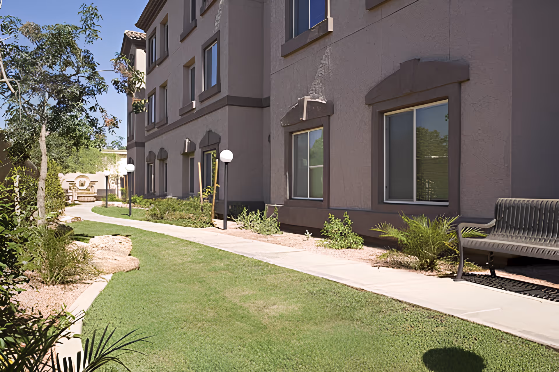 Outdoor walkway alongside a multi-story building with windows, surrounded by green grass, plants, trees, and a bench. The sky is clear and blue.
