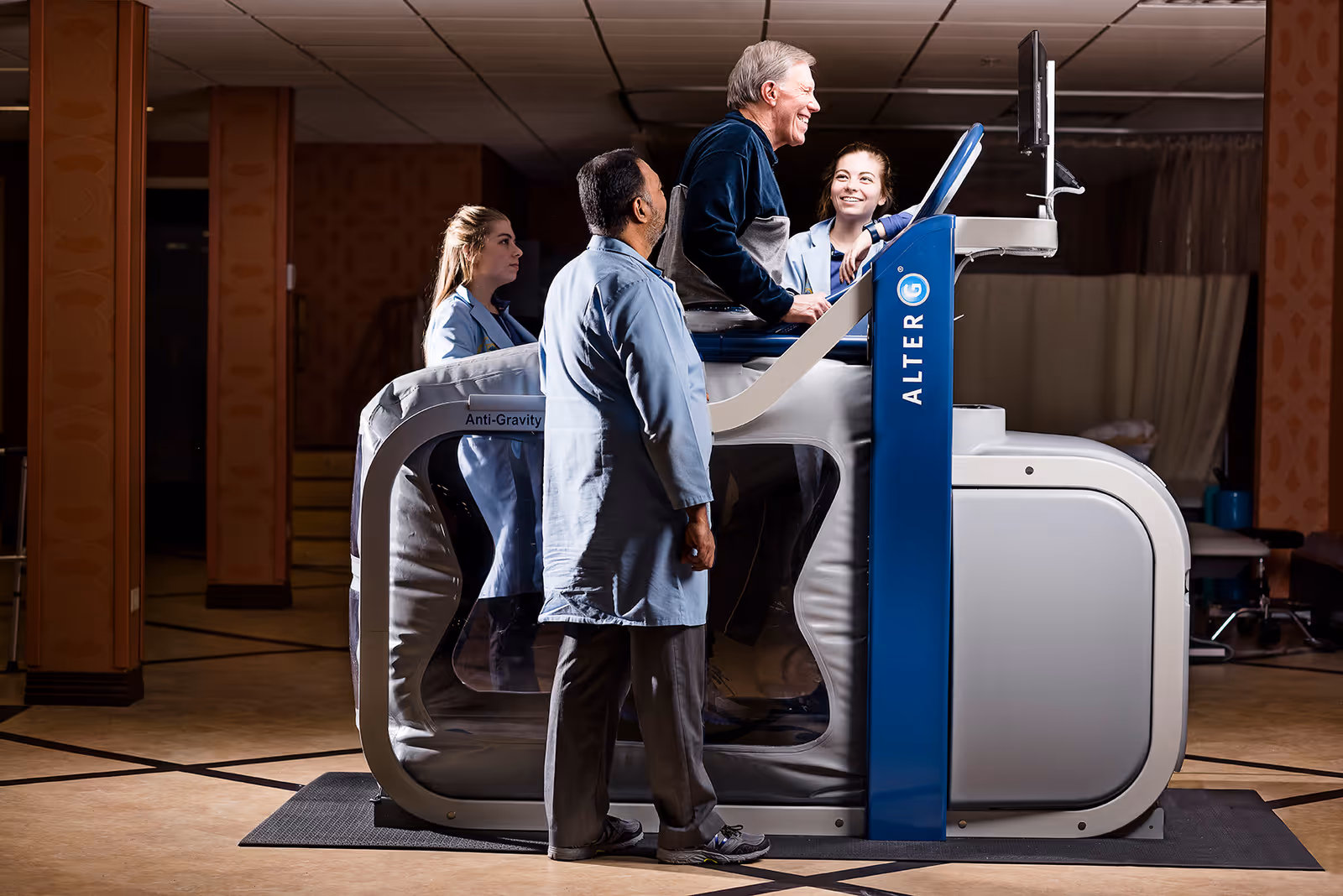 An elderly man is using an AlterG anti-gravity treadmill while two healthcare professionals observe and assist him in a clinical setting.