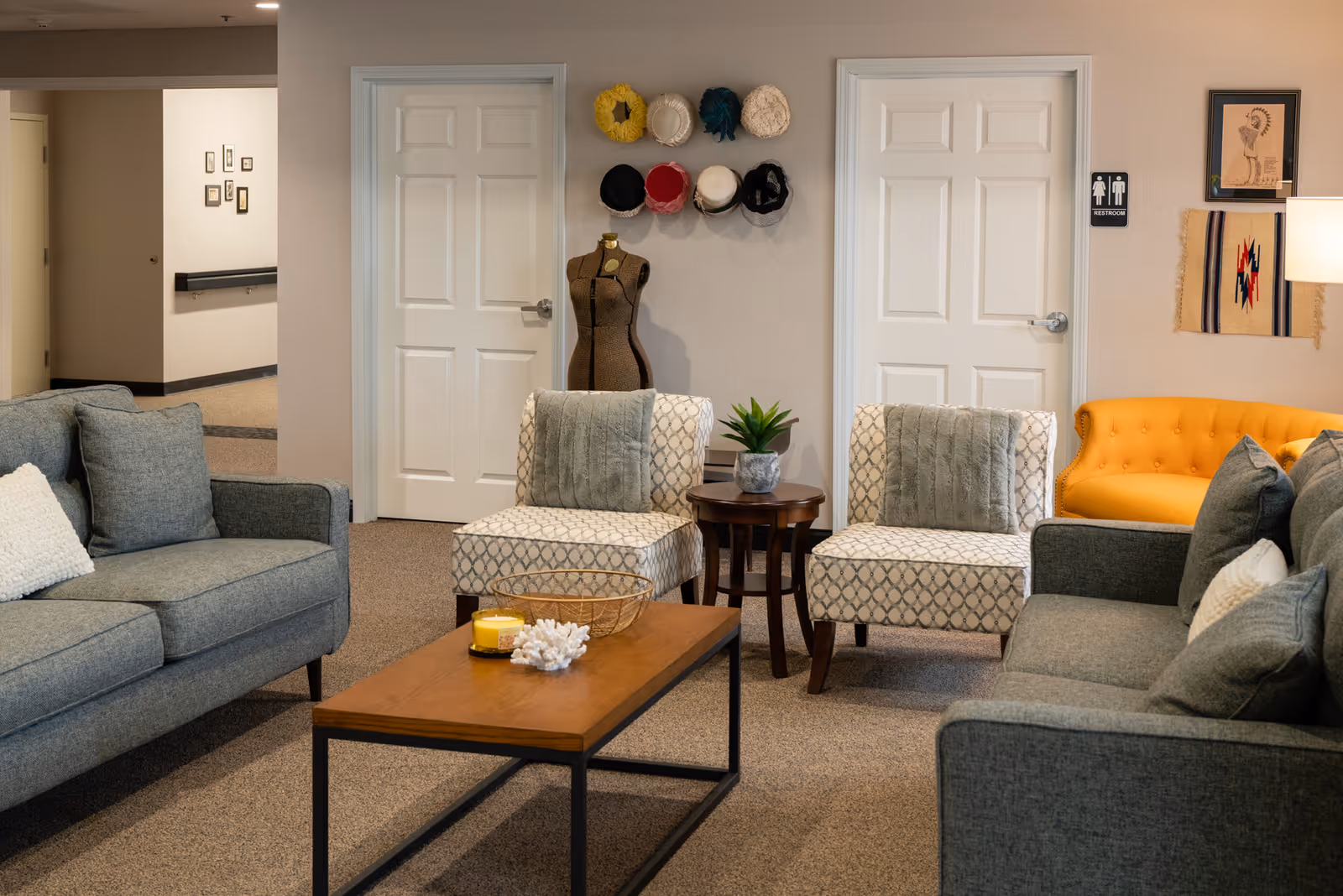 Cozy lounge area with gray sofas, patterned chairs, a wooden coffee table, and a dress form under hats mounted on the wall.