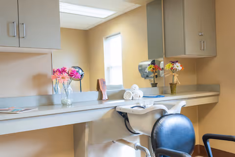 A salon-style hair-washing station with a reclining chair, shampoo sink, countertop, mirror, cabinets and vases of flowers.