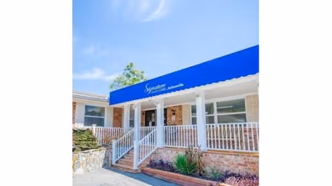 Front entrance of a brick nursing center with a blue awning, white railing, and steps leading to double doors.