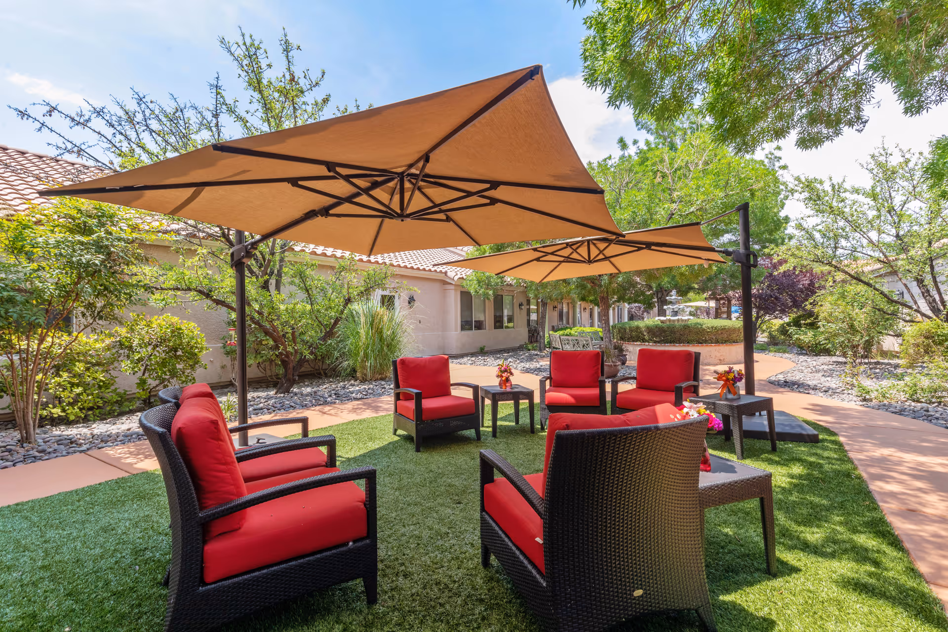 Courtyard with red-cushioned wicker lounge chairs grouped under large beige umbrellas on a grassy patio beside a building.