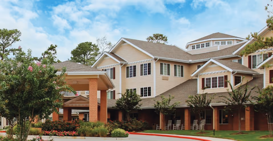 Exterior view of a multi-story senior living facility with beige siding and brick accents, surrounded by landscaped greenery and trees under a partly cloudy sky.