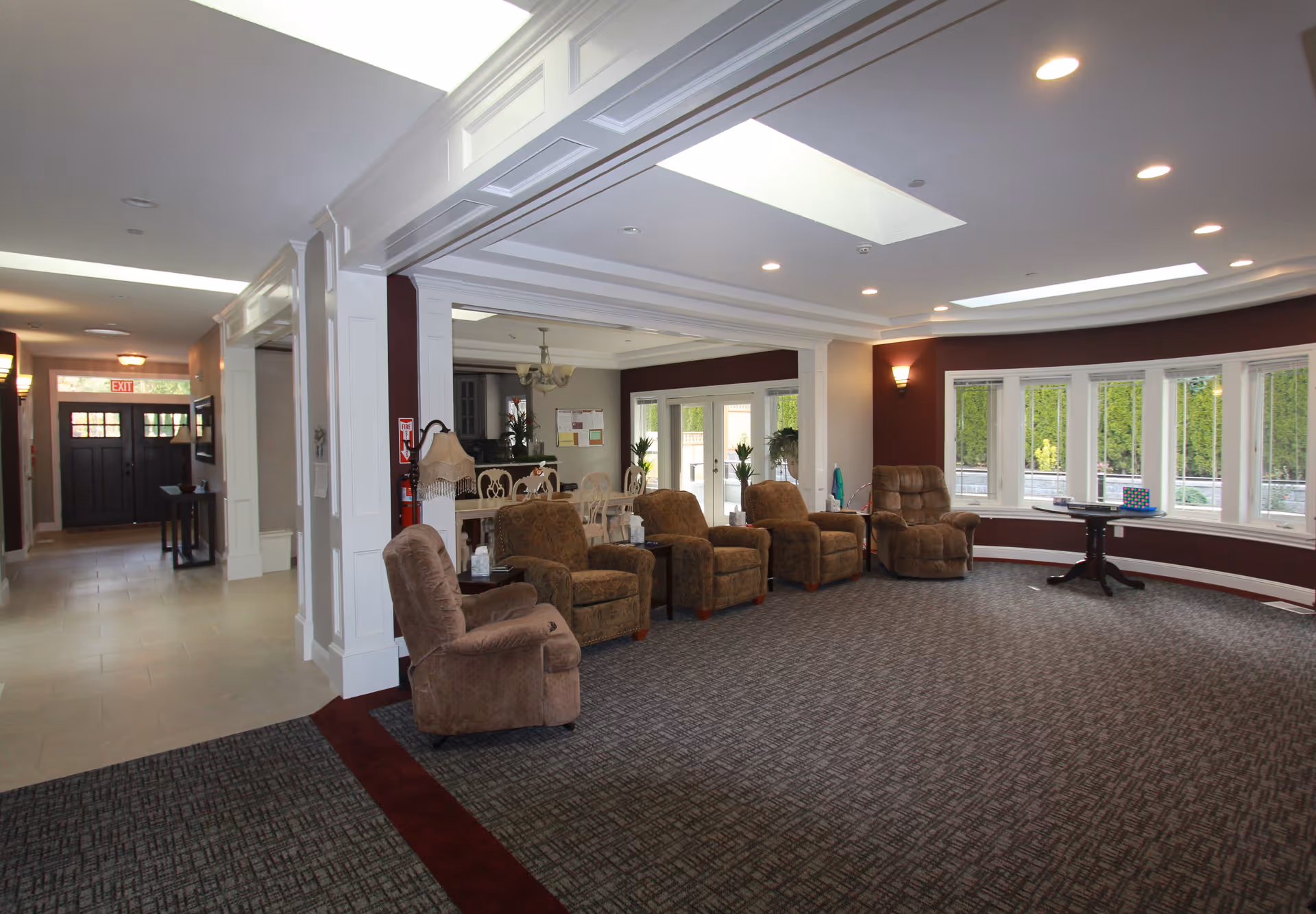 A spacious common area in a senior care facility with several brown upholstered armchairs arranged in a row facing a large window. The room has a patterned carpet, white walls with maroon accents, recessed lighting, and a skylight. In the background, there is a dining area with a table and chairs, and a hallway leading to a dark wooden door with an exit sign above it.