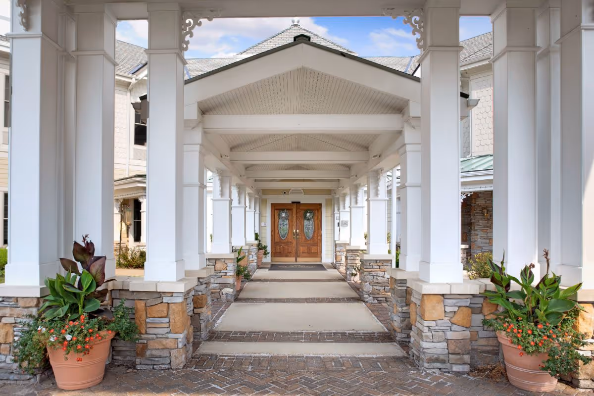 Covered columned entrance walkway with stone bases and potted plants leading to double wooden doors.