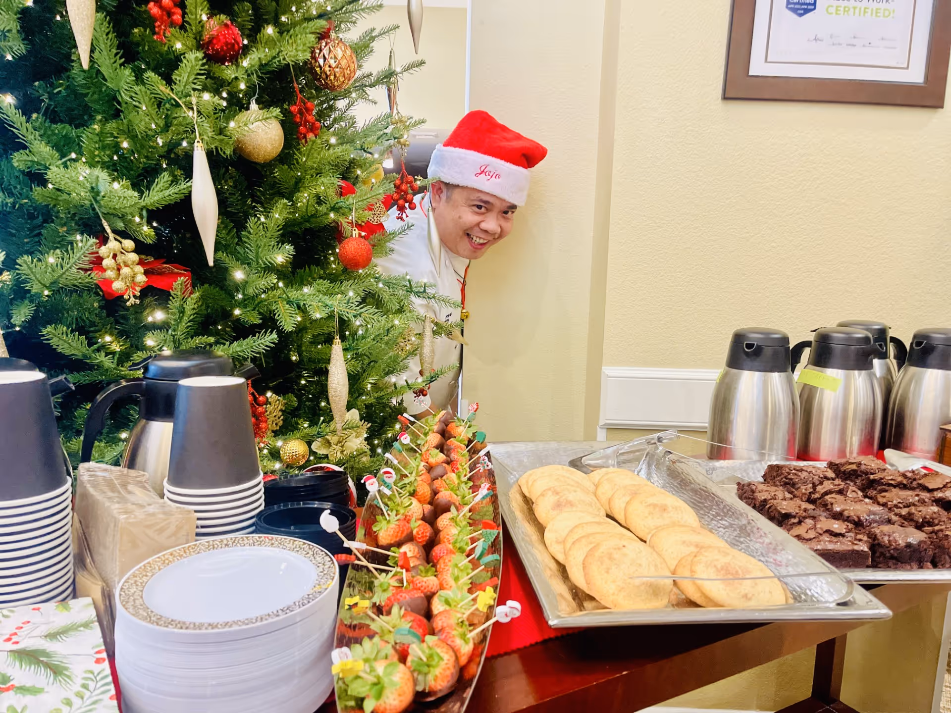 Holiday buffet table with plates, coffee carafes, cookies, brownies and chocolate-covered strawberries beside a decorated Christmas tree and a staff member in a Santa hat peeking out.