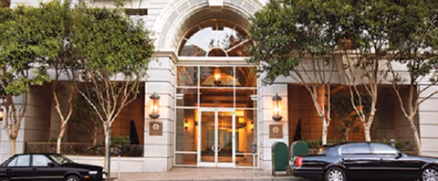 Stone-clad building entrance with an arched glass doorway flanked by trees and parked cars on the street.