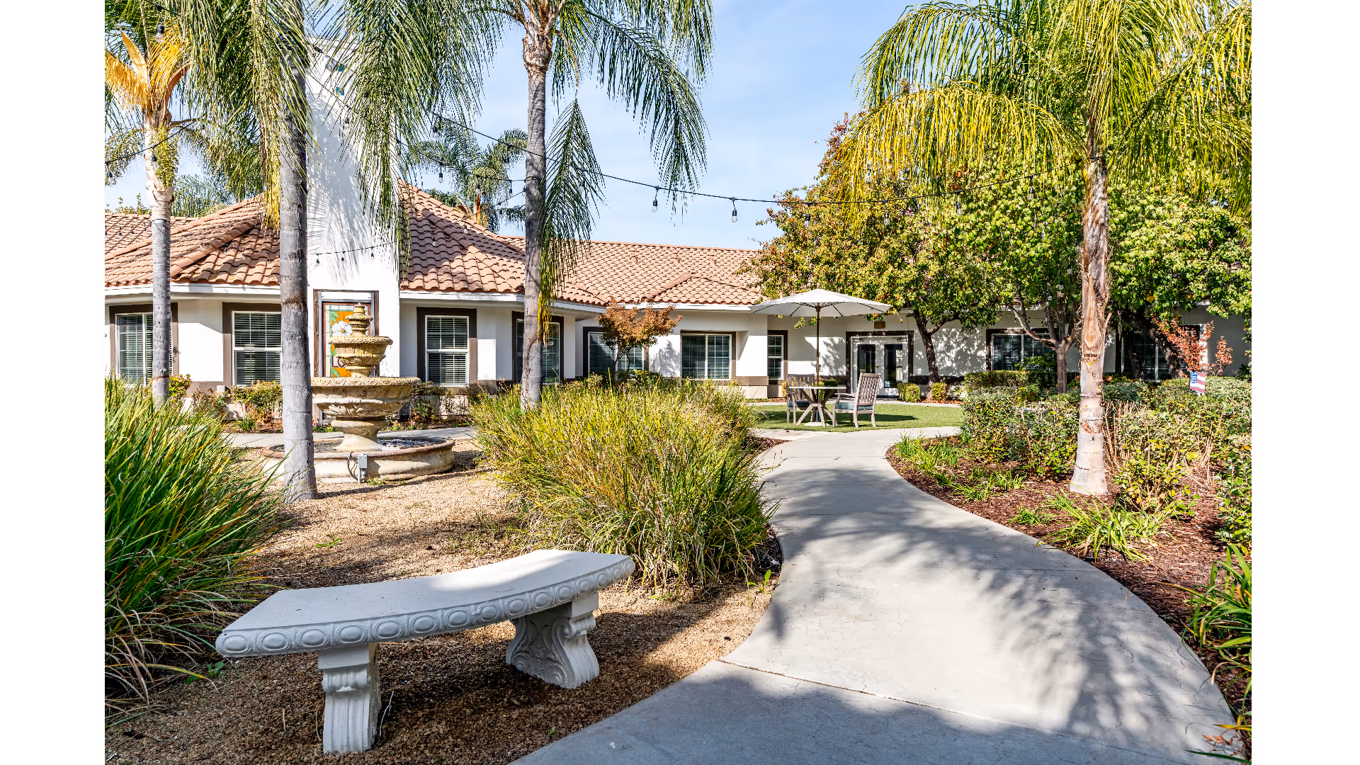 Outdoor garden area at Westmont of Fresno featuring a curved concrete pathway, a stone bench, palm trees, a multi-tiered fountain, and patio furniture with an umbrella. The building with a tiled roof and windows is visible in the background under a clear blue sky.