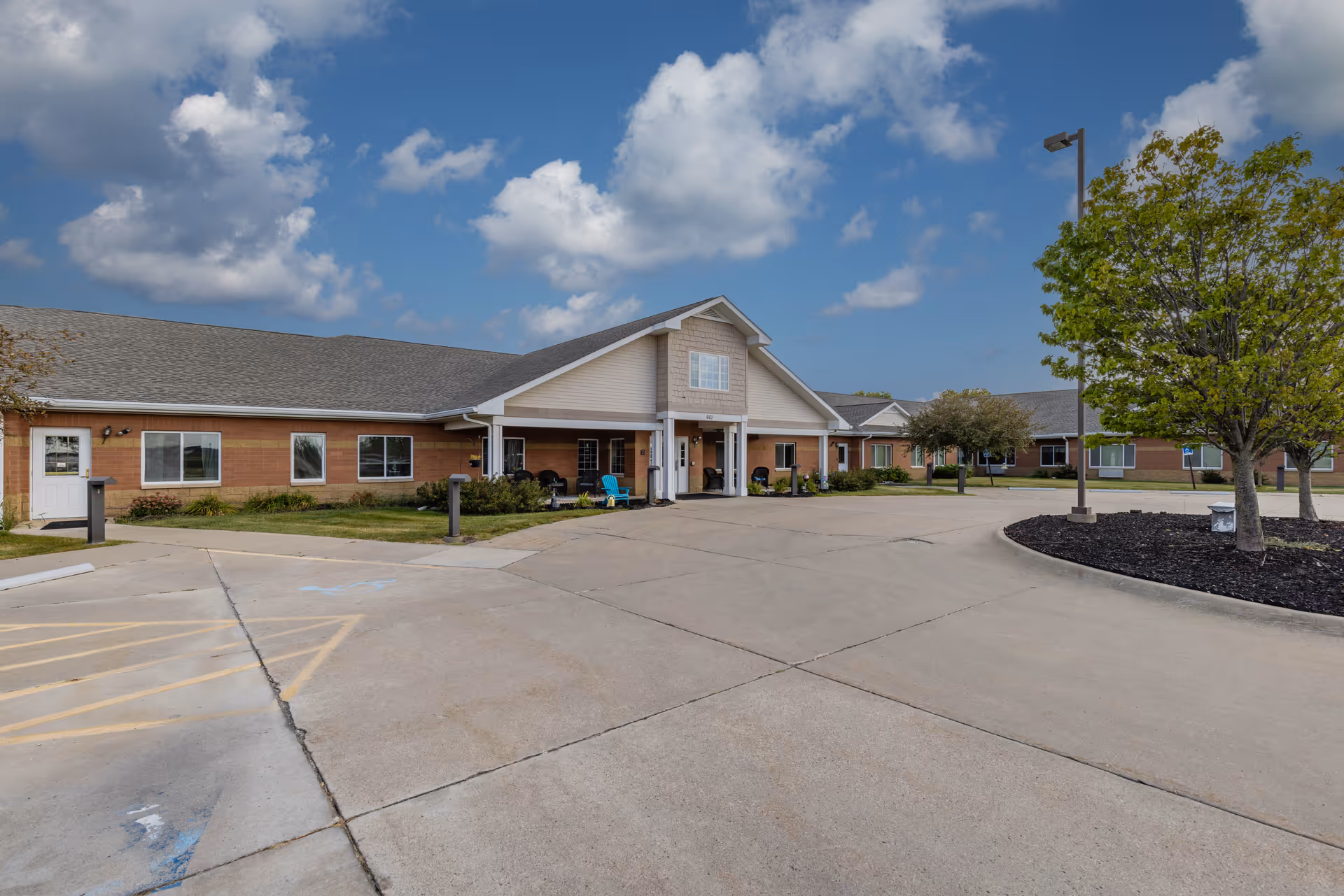 Front entrance of a single-story brick senior living building with a covered portico, driveway, and landscaped trees under a partly cloudy sky.