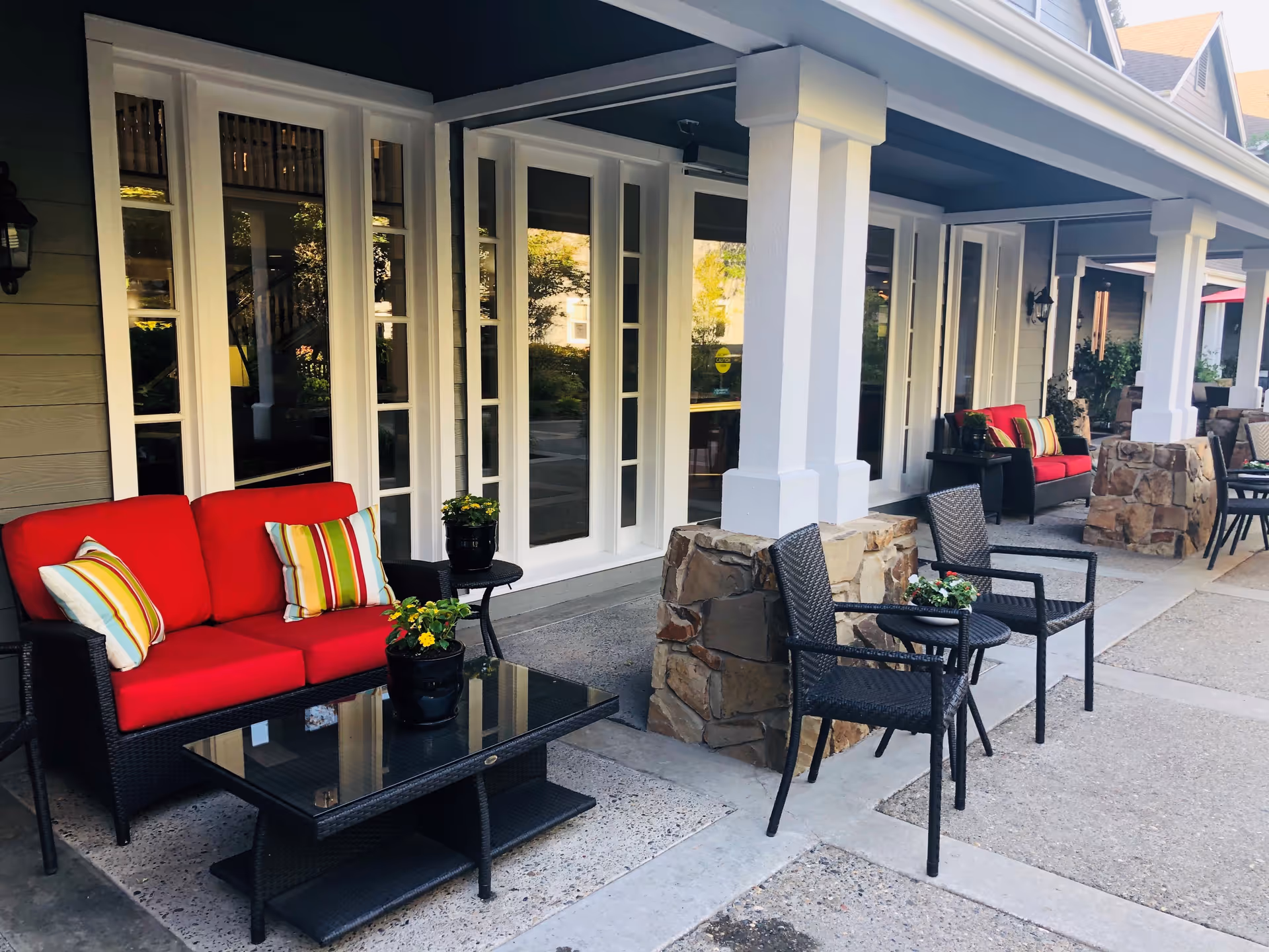 Outdoor patio area at Aegis of Carmichael featuring black wicker furniture with red cushions and striped pillows, black wicker chairs, glass-top coffee tables, potted plants, and white pillars with stone bases along the building facade.