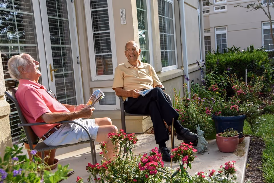 Two elderly men sitting and smiling while reading newspapers on chairs outside a building, surrounded by colorful flowers and greenery in a garden area.