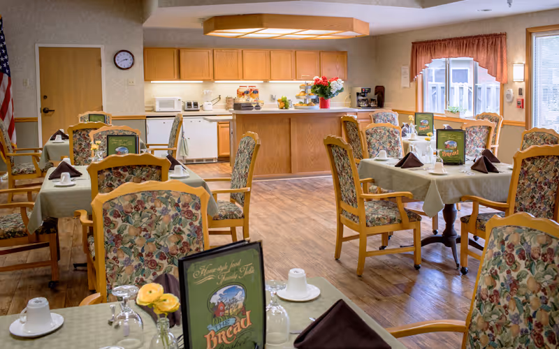 A dining room with several tables covered in light green tablecloths, each set with white cups, glasses, and brown napkins. The chairs have wooden frames with floral-patterned upholstery. In the background, there is a kitchen area with wooden cabinets, a countertop, and various kitchen appliances. A window with a red valance lets in natural light.